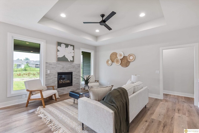 living room featuring ceiling fan, light wood-type flooring, a tray ceiling, and a tiled fireplace