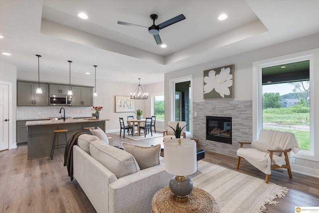 living room featuring hardwood / wood-style floors, a raised ceiling, a fireplace, ceiling fan with notable chandelier, and sink