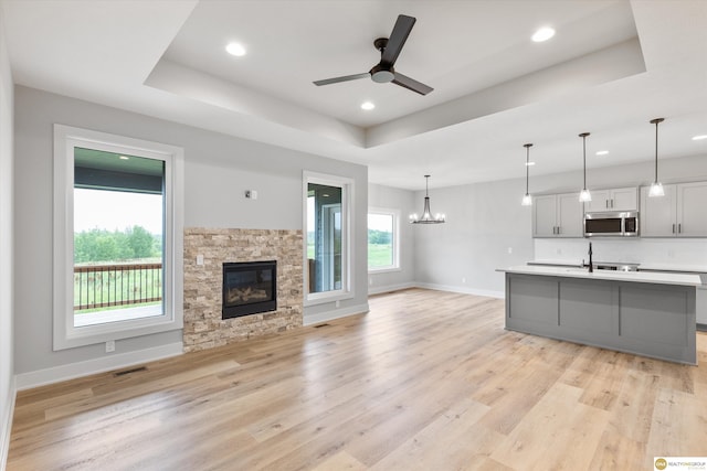 unfurnished living room with ceiling fan with notable chandelier, a tray ceiling, light hardwood / wood-style flooring, and a fireplace