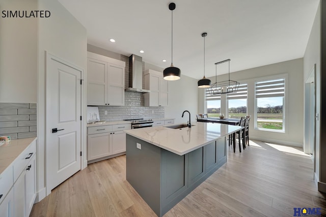 kitchen featuring a center island with sink, wall chimney exhaust hood, white cabinets, light stone counters, and sink