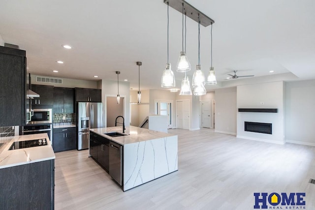 kitchen featuring an island with sink, appliances with stainless steel finishes, decorative light fixtures, light stone counters, and sink