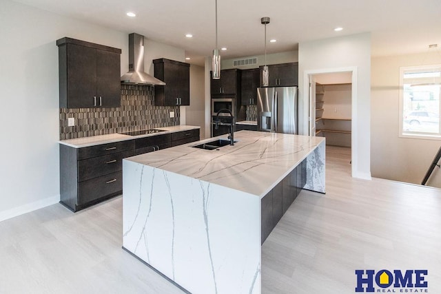 kitchen featuring stainless steel refrigerator with ice dispenser, a kitchen island with sink, hanging light fixtures, wall chimney range hood, and sink