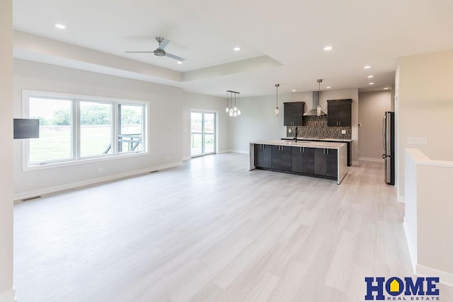 kitchen featuring wall chimney range hood, a center island with sink, decorative backsplash, light wood-type flooring, and stainless steel fridge