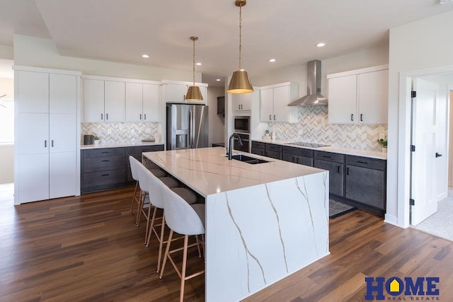 kitchen featuring a center island with sink, stainless steel refrigerator with ice dispenser, sink, white cabinetry, and wall chimney exhaust hood