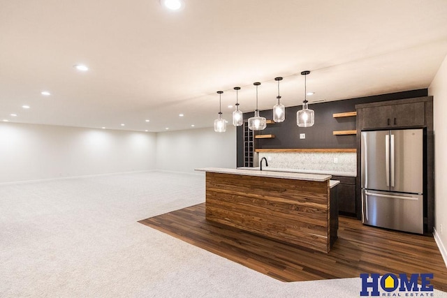 kitchen featuring a kitchen island with sink, dark brown cabinets, stainless steel refrigerator, and decorative backsplash