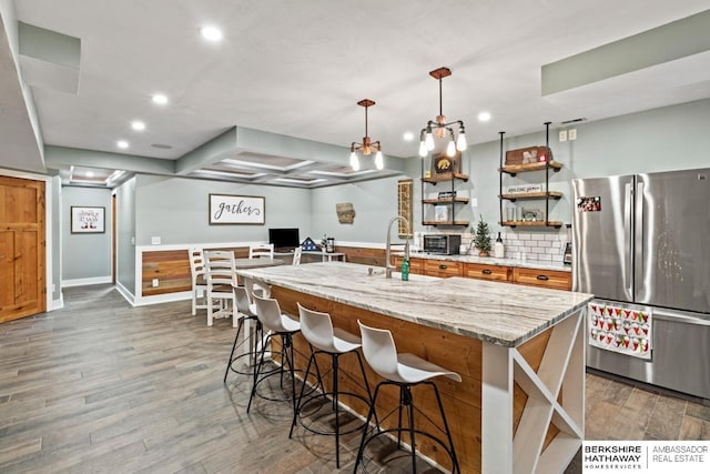 kitchen featuring dark hardwood / wood-style flooring, decorative light fixtures, light stone counters, an island with sink, and stainless steel refrigerator