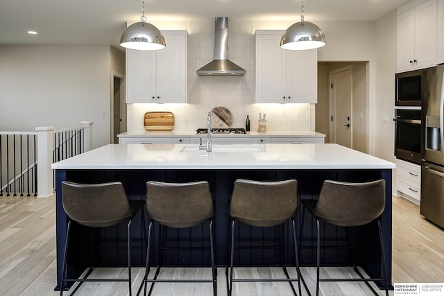 kitchen featuring a center island with sink, white cabinetry, appliances with stainless steel finishes, and wall chimney range hood