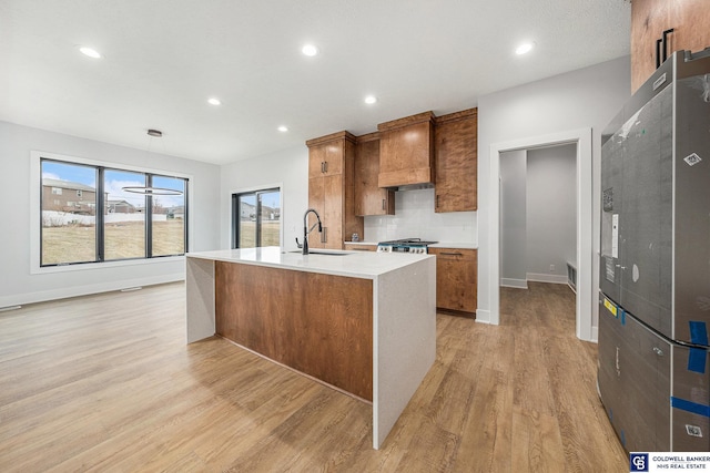 kitchen with sink, a center island with sink, custom range hood, light hardwood / wood-style floors, and backsplash