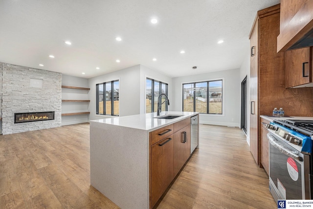 kitchen featuring a stone fireplace, sink, a center island with sink, light wood-type flooring, and appliances with stainless steel finishes