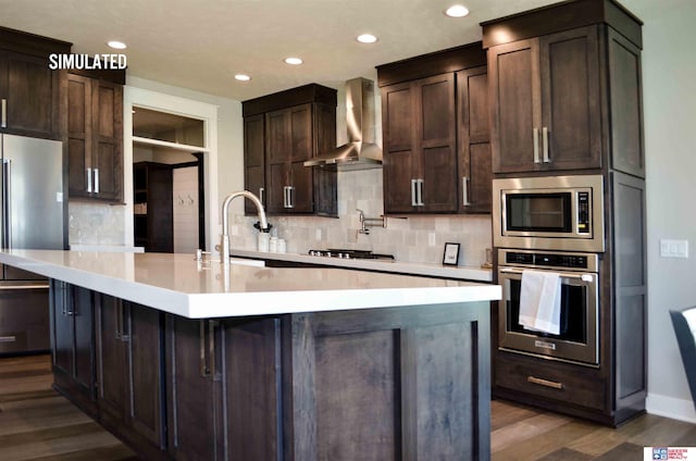 kitchen with an island with sink, appliances with stainless steel finishes, sink, and wall chimney range hood