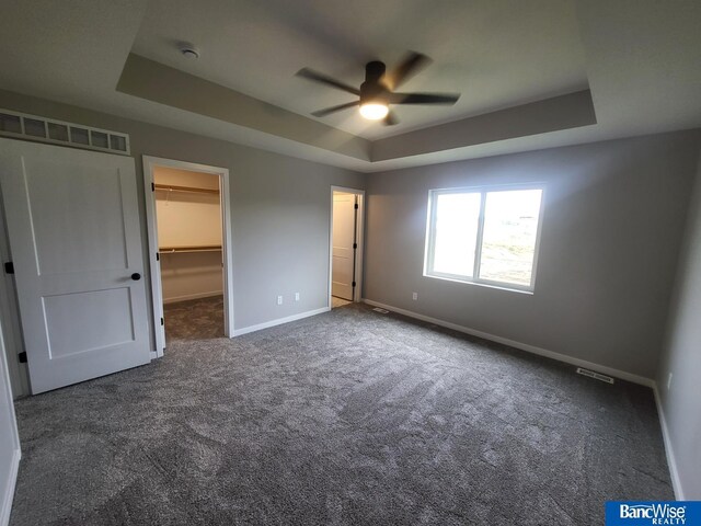 unfurnished bedroom with a spacious closet, dark carpet, ceiling fan, a raised ceiling, and a closet