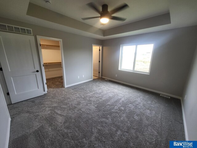 unfurnished bedroom featuring ceiling fan, a tray ceiling, dark carpet, and a spacious closet