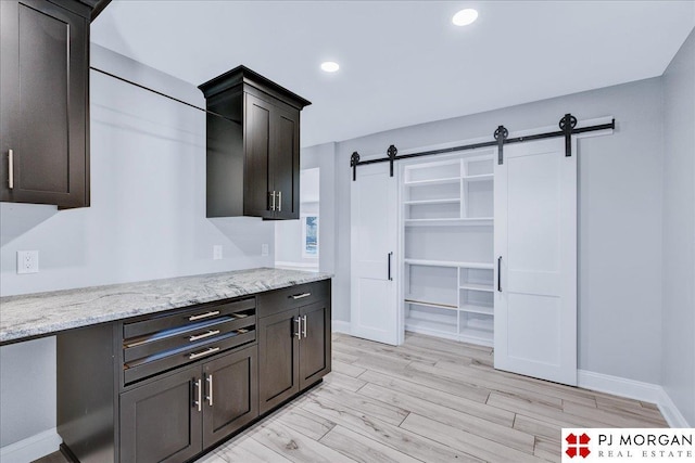 kitchen featuring dark brown cabinetry, a barn door, light wood-type flooring, and light stone counters