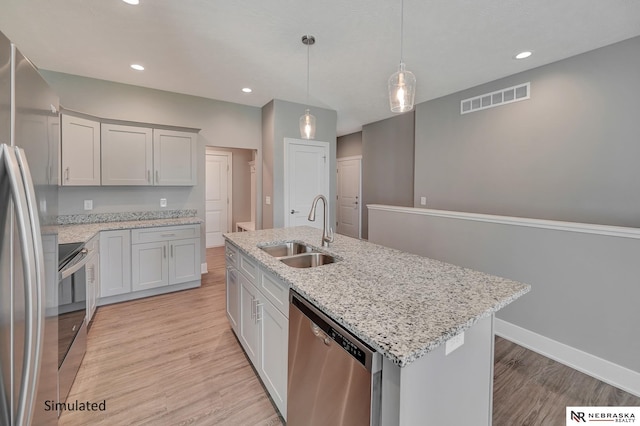 kitchen with sink, appliances with stainless steel finishes, an island with sink, pendant lighting, and light stone countertops