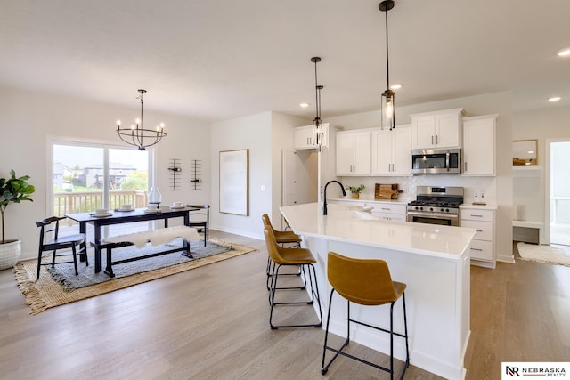 kitchen with sink, appliances with stainless steel finishes, a kitchen island with sink, white cabinets, and decorative light fixtures