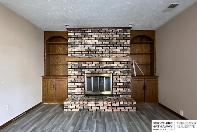 unfurnished living room featuring built in shelves, dark wood-type flooring, a brick fireplace, and a textured ceiling