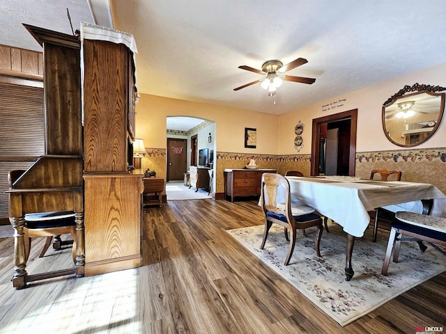 dining area featuring arched walkways, wainscoting, dark wood-style floors, ceiling fan, and a textured ceiling