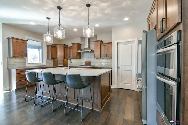 kitchen with dark hardwood / wood-style floors, an island with sink, hanging light fixtures, stainless steel appliances, and wall chimney exhaust hood