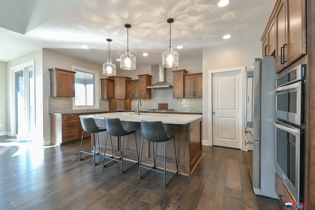 kitchen with wall chimney range hood, appliances with stainless steel finishes, a kitchen island with sink, hanging light fixtures, and dark hardwood / wood-style flooring