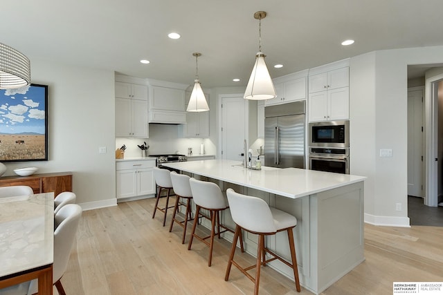 kitchen with white cabinetry, hanging light fixtures, built in appliances, light hardwood / wood-style floors, and an island with sink