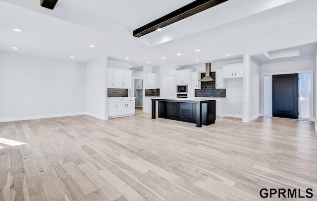 kitchen featuring an island with sink, white cabinets, decorative backsplash, wall chimney exhaust hood, and light hardwood / wood-style flooring