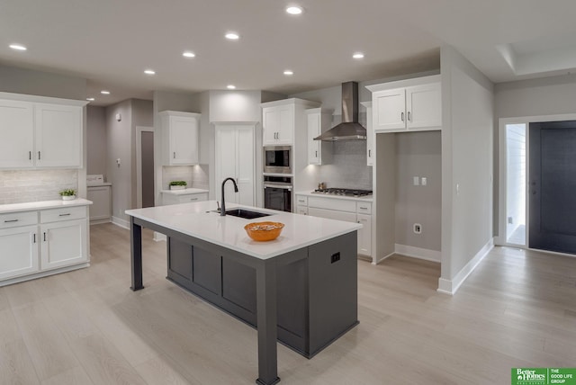 kitchen with wall chimney exhaust hood, sink, white cabinetry, stainless steel appliances, and a kitchen island with sink