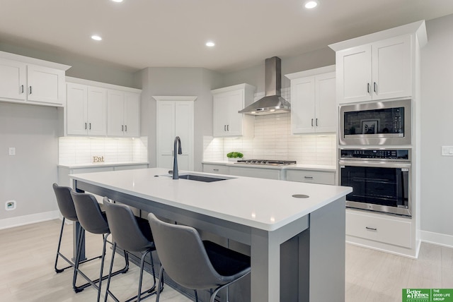 kitchen with sink, appliances with stainless steel finishes, wall chimney range hood, a kitchen island with sink, and white cabinets