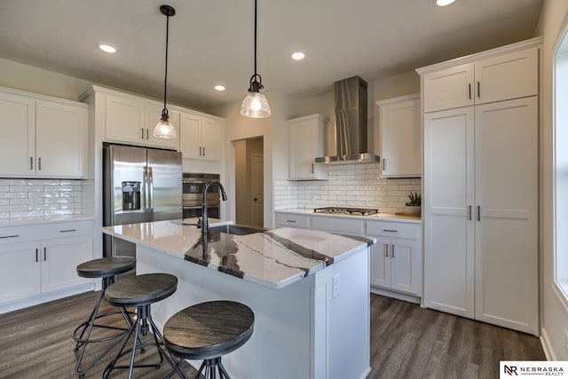 kitchen featuring wall chimney exhaust hood, sink, hanging light fixtures, an island with sink, and white cabinets