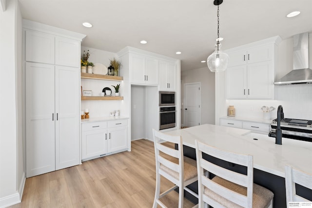 kitchen featuring wall chimney exhaust hood, decorative light fixtures, stainless steel appliances, light hardwood / wood-style floors, and white cabinets