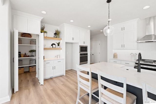 kitchen featuring hanging light fixtures, light wood-type flooring, appliances with stainless steel finishes, wall chimney range hood, and white cabinets