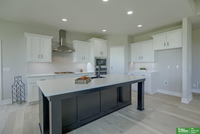 kitchen with white cabinets, stainless steel appliances, an island with sink, and wall chimney range hood