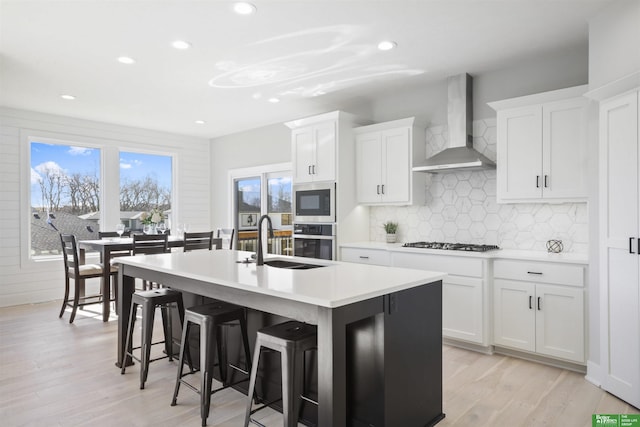 kitchen with light countertops, white cabinets, a sink, wall chimney range hood, and an island with sink