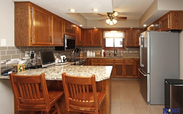 kitchen with brown cabinets, stainless steel appliances, a sink, light stone countertops, and a peninsula