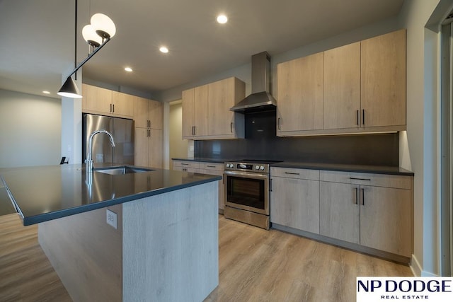 kitchen featuring wall chimney range hood, stainless steel appliances, an island with sink, decorative light fixtures, and light brown cabinets