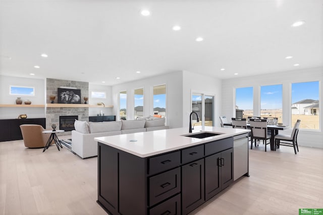 kitchen with sink, a kitchen island with sink, light hardwood / wood-style floors, a stone fireplace, and stainless steel dishwasher