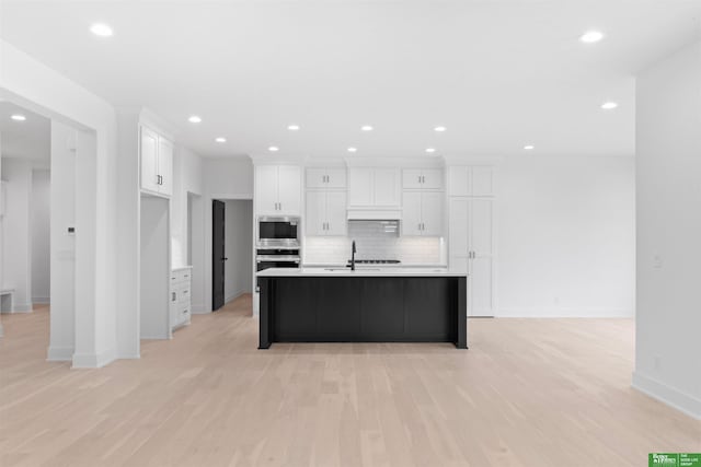 kitchen with white cabinets, backsplash, a kitchen island with sink, stainless steel appliances, and light wood-type flooring