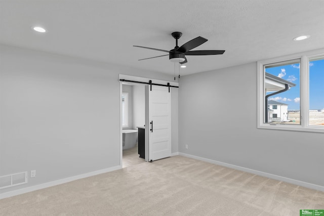 unfurnished bedroom featuring ensuite bath, light colored carpet, a barn door, and ceiling fan