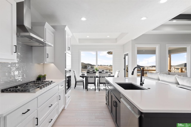 kitchen with sink, a raised ceiling, stainless steel appliances, a kitchen island with sink, and wall chimney range hood