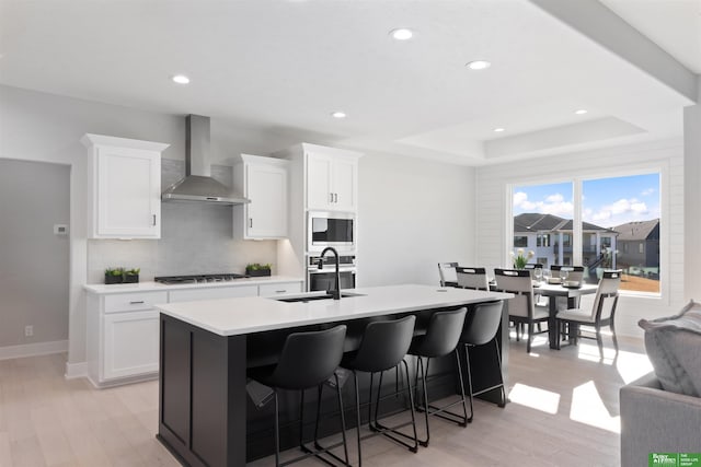 kitchen with sink, white cabinetry, a tray ceiling, an island with sink, and wall chimney exhaust hood