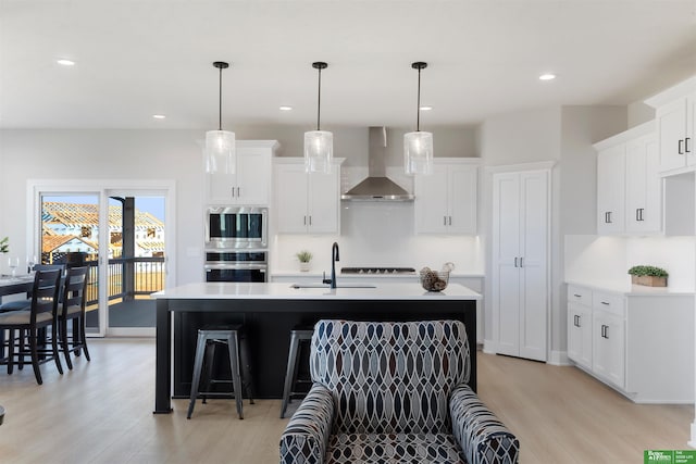 kitchen with white cabinetry, stainless steel appliances, wall chimney exhaust hood, and a center island with sink