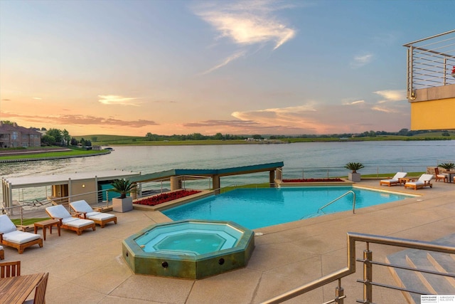 pool at dusk with an in ground hot tub, a water view, and a patio