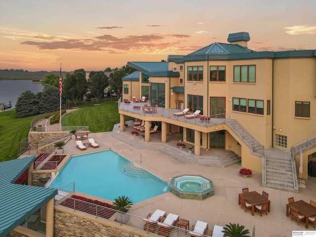 pool at dusk with a patio area, a lawn, a water view, and an in ground hot tub