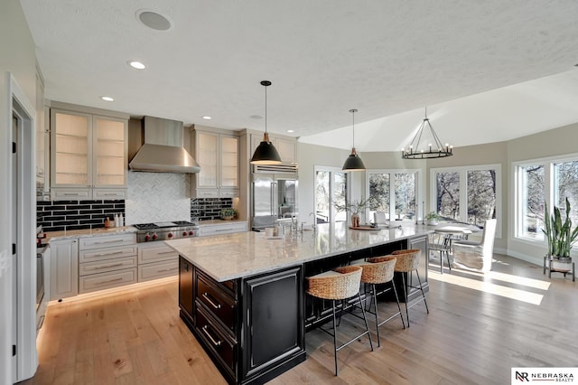 kitchen featuring wall chimney exhaust hood, light stone counters, a large island with sink, pendant lighting, and stainless steel appliances