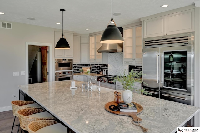 kitchen featuring a large island, backsplash, stainless steel appliances, light stone counters, and wall chimney exhaust hood