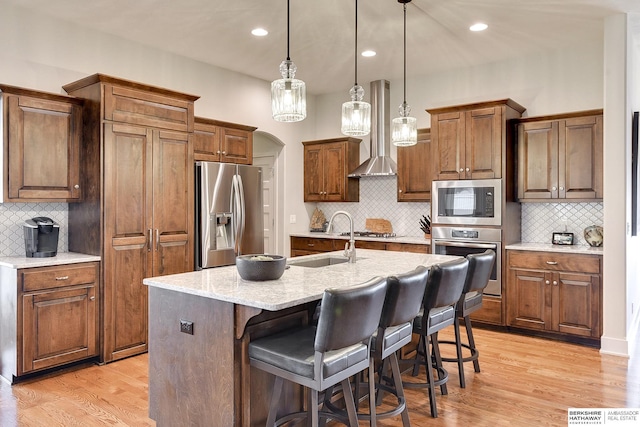 kitchen featuring pendant lighting, sink, wall chimney range hood, appliances with stainless steel finishes, and a kitchen island with sink