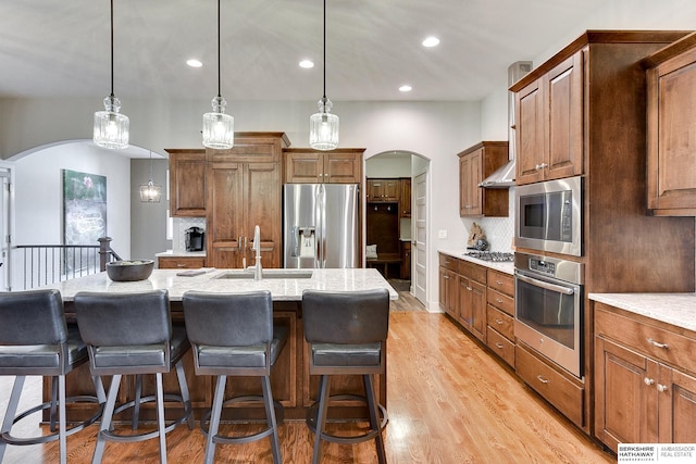 kitchen with sink, a kitchen island with sink, stainless steel appliances, light stone counters, and decorative light fixtures