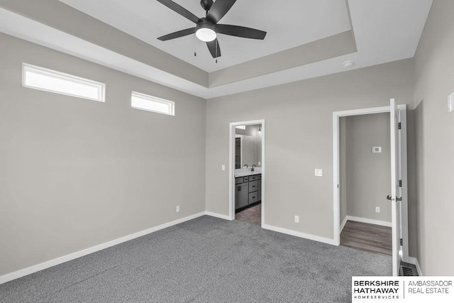 unfurnished bedroom featuring a raised ceiling, dark colored carpet, connected bathroom, and ceiling fan