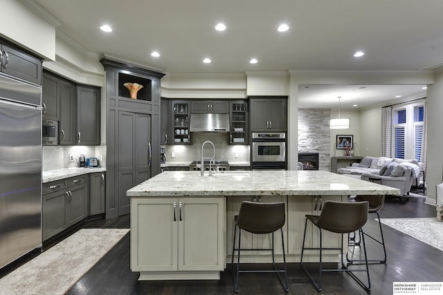 kitchen featuring gray cabinetry, built in appliances, a kitchen breakfast bar, an island with sink, and light stone countertops