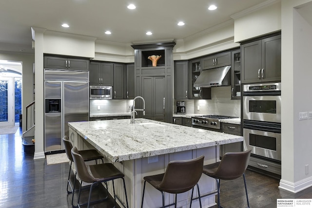 kitchen with an island with sink, a kitchen breakfast bar, ornamental molding, built in appliances, and light stone countertops