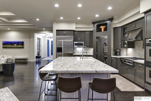 kitchen featuring an island with sink, light stone countertops, built in appliances, and a kitchen bar